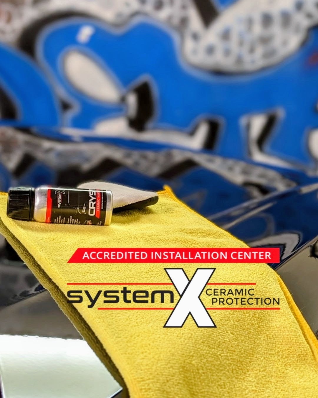 A bottle of System X Crystal coating on a yellow microfiber towel laid over a freshly coated hood, marked System X Ceramic Protection Accredited Installation Center